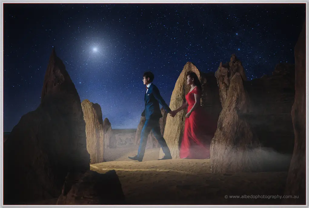 Couple in a red gown and navy suit walk hand in hand among the Pinnacles under a starlit desert night