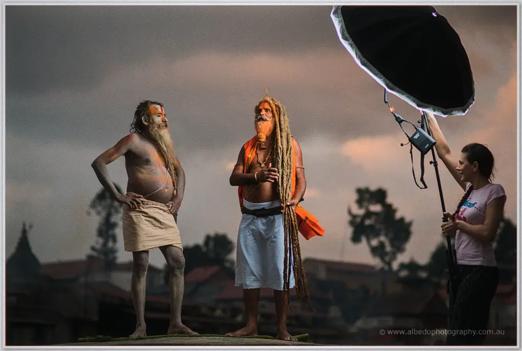 Two sadhus stand for a portrait at dusk while an assistant holds a studio umbrella beside them in Nepal