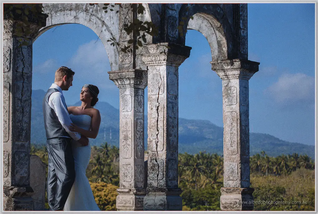 Bride and groom embrace among weathered stone arches above palm groves and distant blue mountains