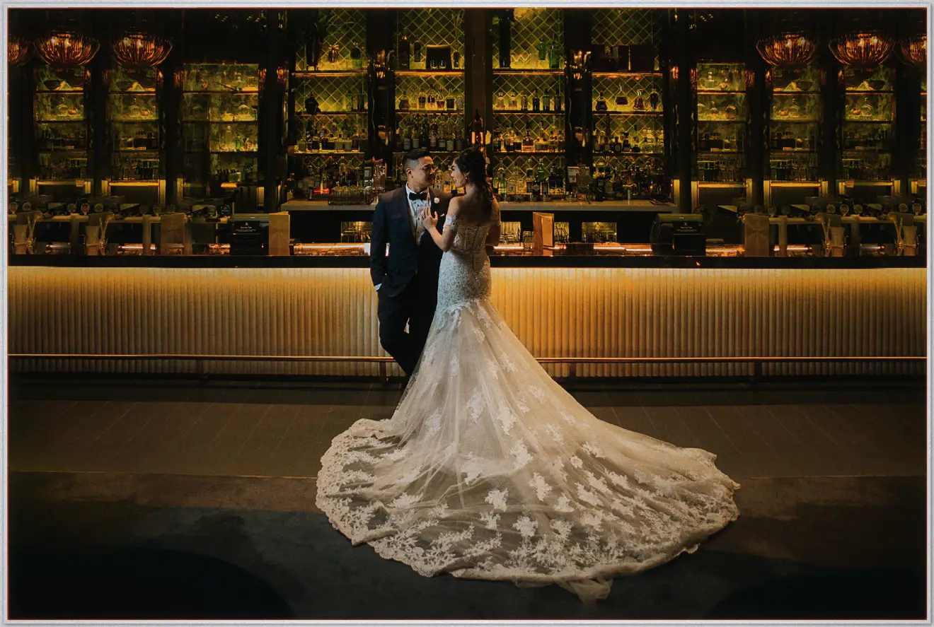 Denis and Suzii lean together by a golden-lit bar, her wide lace train pooled across the floor