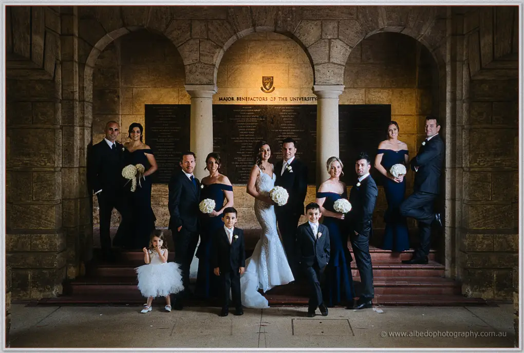 Crystal and Joe's bridal party in navy gowns and dark suits posed on sunlit stone steps beneath arches