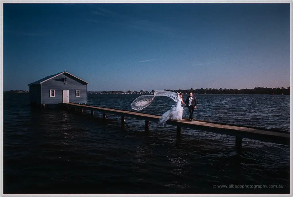 Tracy and Leandro walk the timber jetty past the still blue boatshed, her veil lifting on the breeze