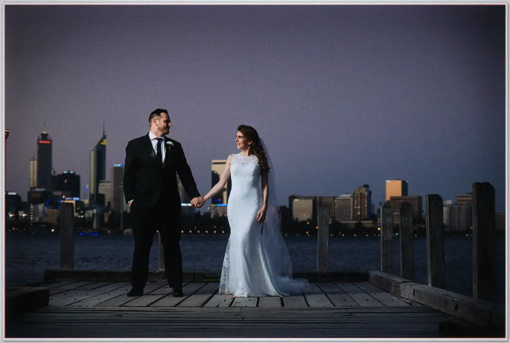Ciara and Josh hold hands on a jetty as city towers glow across the purple twilight water