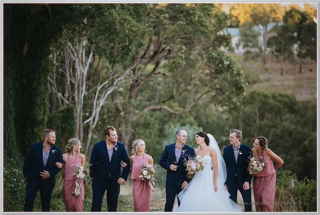 Lucy and Blake laugh with pink-gowned bridesmaids and groomsmen among the trees at Darlington Estate
