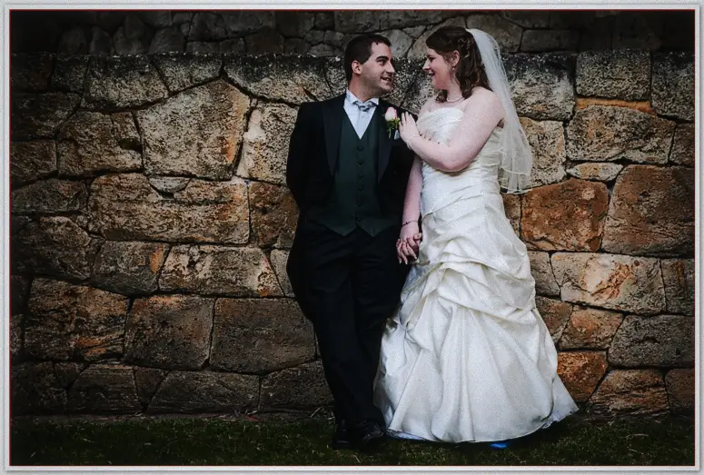 Perth Wedding Photographer - Jaimi and kevin's Wedding Groom in a dark suit and veiled bride lean together against a rough limestone wall in moody light