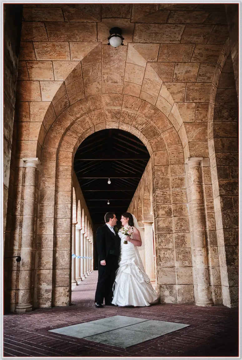 Perth Wedding Photographer - Jaimi and kevin's Wedding Bride and groom embrace beneath the soaring sandstone arches of the university colonnade, bouquet in hand