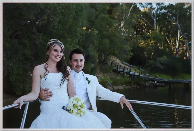 Lauren rests her rose bouquet on Simon's lap aboard the boat bow, golden light on the riverbank