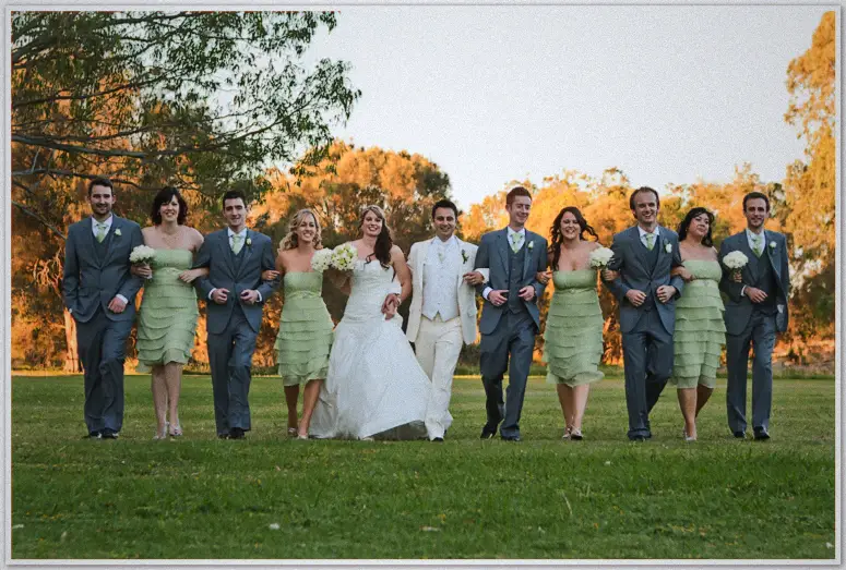 The bridal party walks arm in arm across a sunset-lit park, blue suits and sage dresses glowing