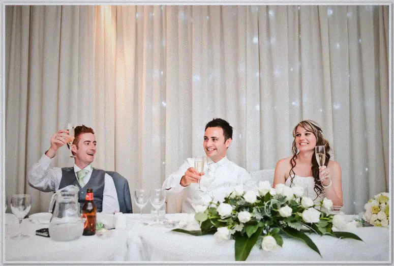 Lauren and Simon toast with champagne beside their grinning best man along a white-rose garland