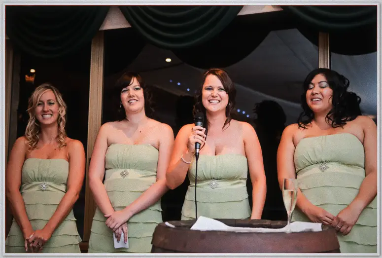 A bridesmaid laughs into the microphone at a wine-barrel podium, three companions smiling behind