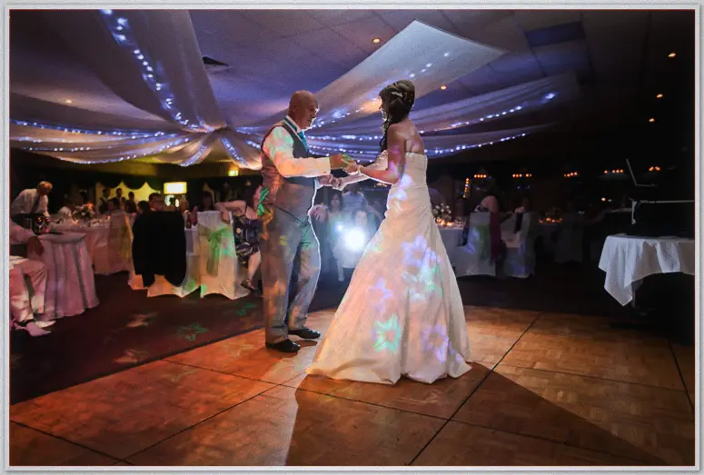 An older gentleman dances with Lauren beneath blue-lit drapes, flower lights pooling on her skirt