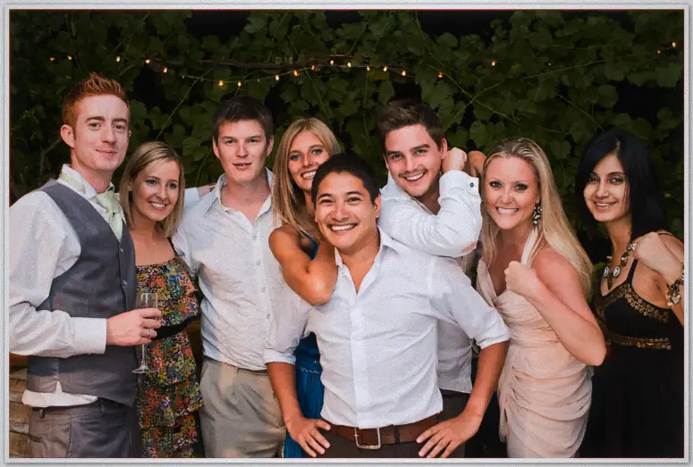 Nine friends in party shirts and dresses strike cheeky poses beneath string lights and vines