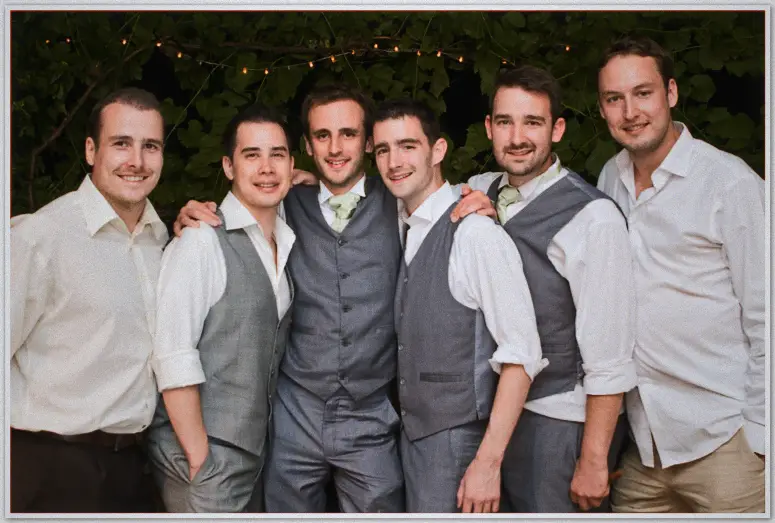 Six groomsmen in grey vests and sage ties stand shoulder to shoulder beneath the vine-covered wall