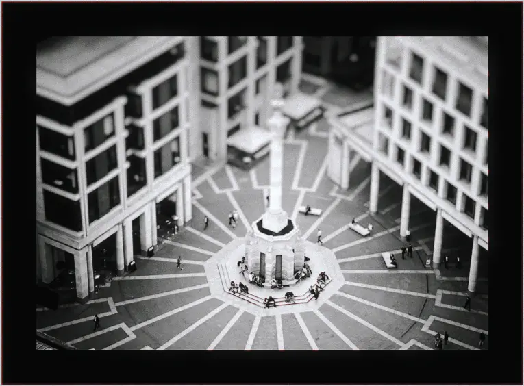 Black and white aerial of workers circling a columned monument in a geometric city square