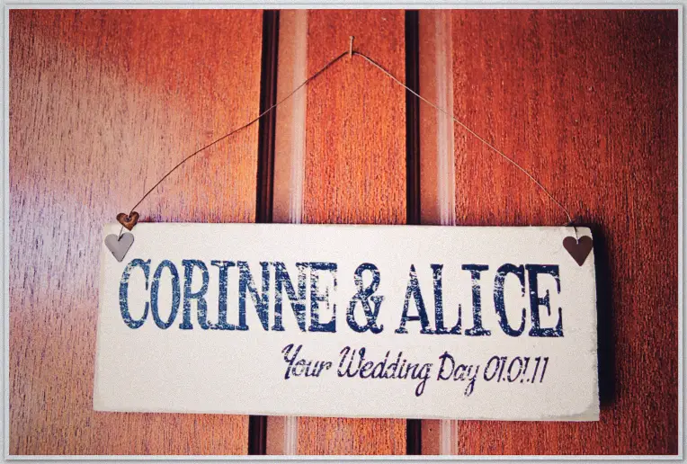 Close-up painted sign reading Corinne & Alice Your Wedding Day with tiny hearts, hung on red timber wall