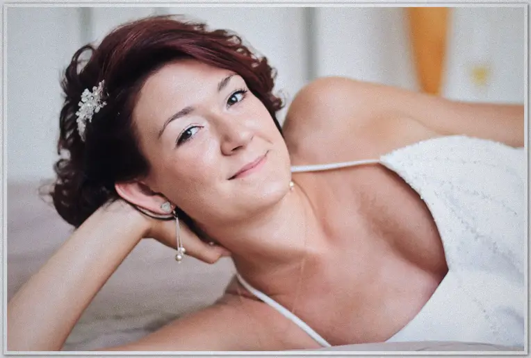 Bride with crystal hairpiece reclines on white bed looking into camera, soft daylight portrait