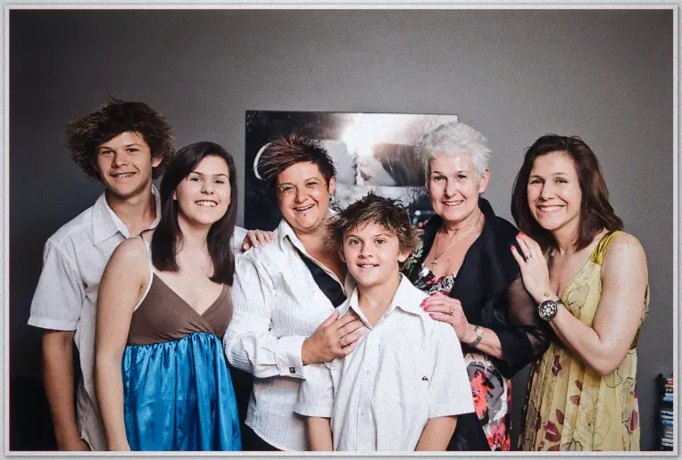 Family of six smiles together on grey lounge wall backdrop, bride-to-be in white shirt at centre