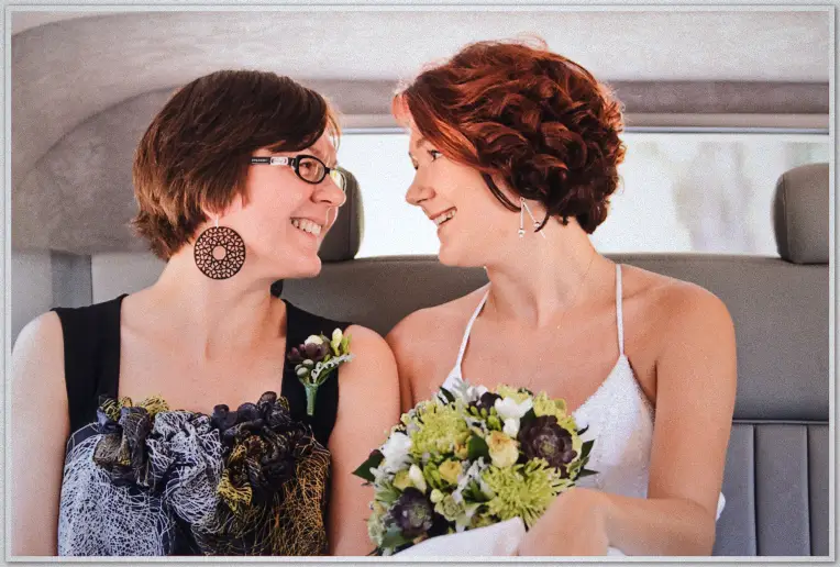 Two brides laugh facing each other in limousine back seat, one holding green and purple bouquet