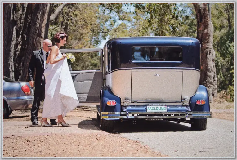Bride steps from vintage navy limousine holding bouquet, father in suit beside her under gum trees