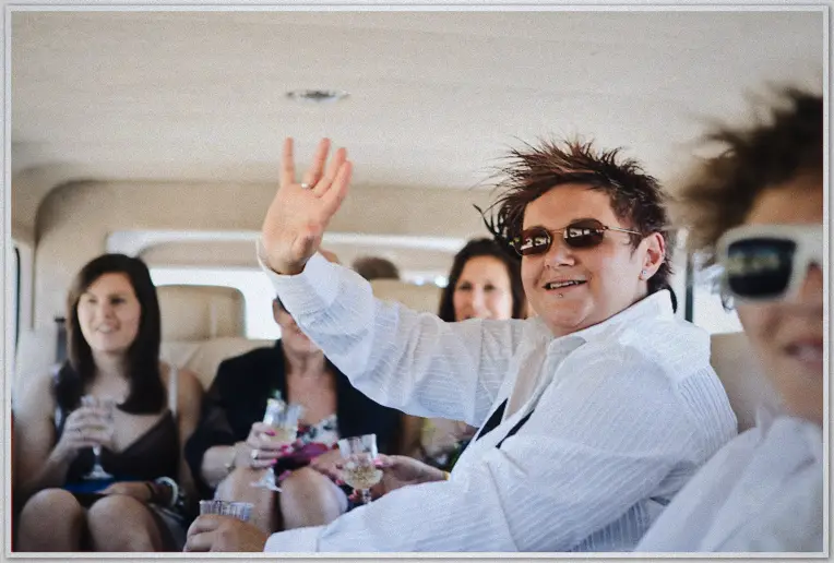Wedding party toasts with champagne in limo, groom-style bride in sunglasses waves at camera