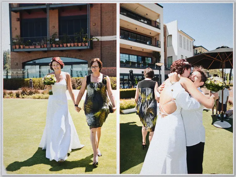 Bride walks hand in hand with guest across riverside lawn, then hugs her partner in sunshine