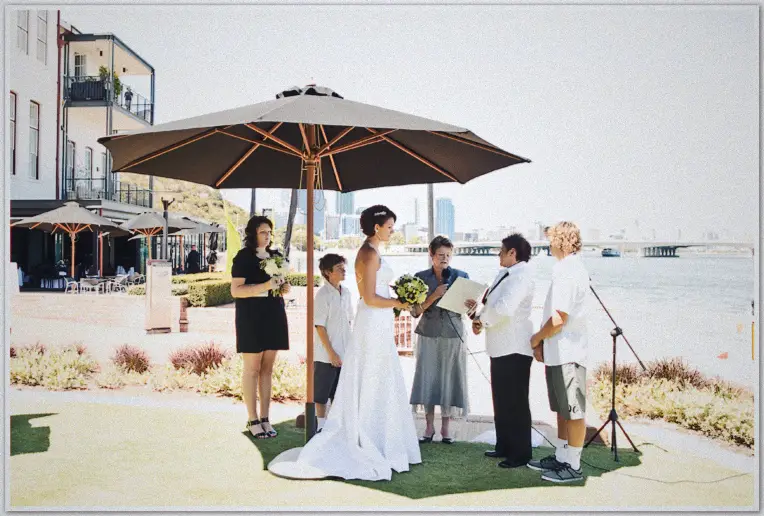 Both brides exchange vows beneath a market umbrella on the river foreshore, city skyline behind