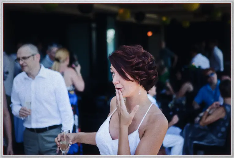 Bride blows a kiss across the reception, champagne in hand, guests mingling in dim warmth