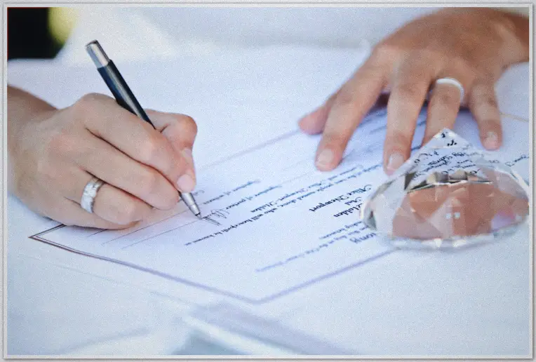 Ringed hand signs the marriage certificate with silver pen, crystal weight holding paper