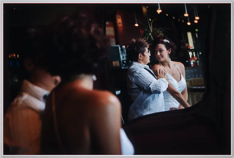 Brides laugh together in the dim lounge, seen reflected in a wide bar mirror at night