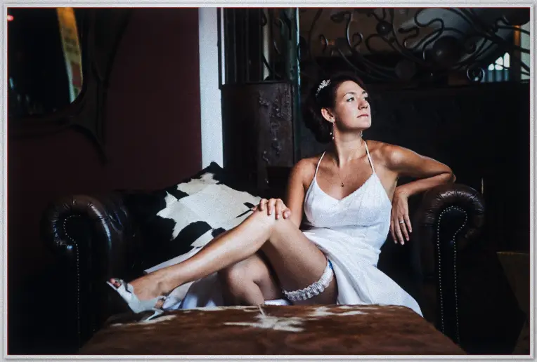 Bride in white gown lounges on a leather chaise beside cowhide rug, garter peeking