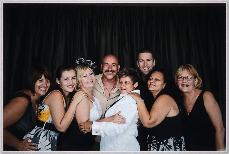 Seven friends squeeze around the bride in a photo booth, arms linked against black drape
