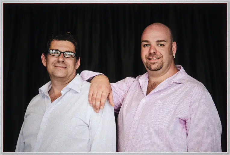 Two men in dress shirts pose arm over shoulder in the photo booth, relaxed grins