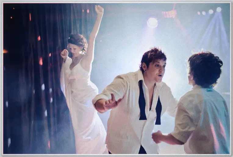 Bride in shirt and tie dances with a boy as her partner raises an arm under hazy reception light