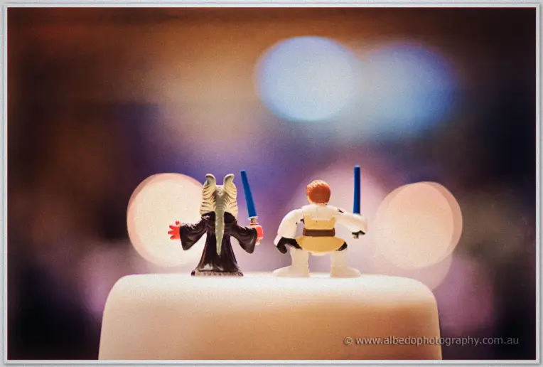 Fremantle Wedding | Charlene and Phil Toy figures wielding lightsabers stand back to back on top of the white wedding cake in dreamy bokeh light