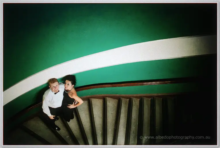Fremantle Wedding | Charlene and Phil Seen from above, Charlene and Phil gaze up from a sweeping spiral staircase against a deep green wall