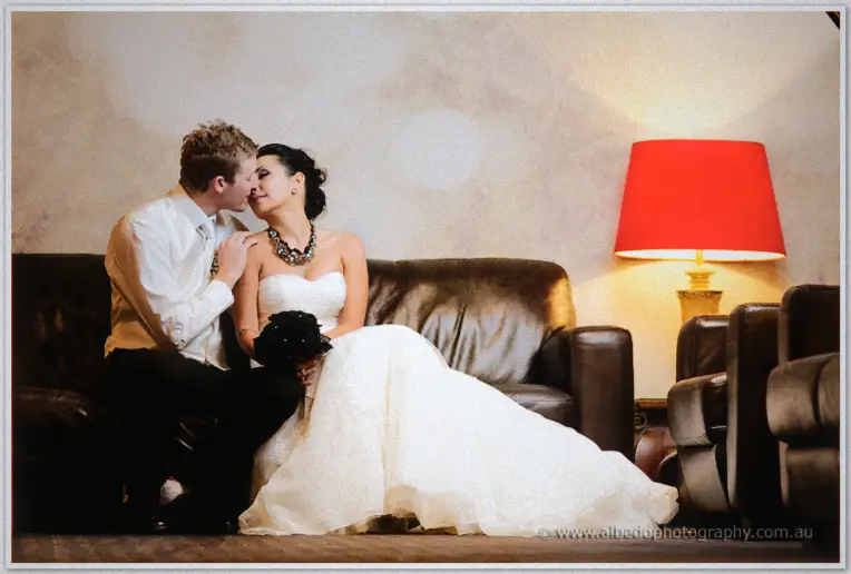 Fremantle Wedding | Charlene and Phil Phil kisses Charlene's cheek on a leather lounge beside a glowing red lampshade