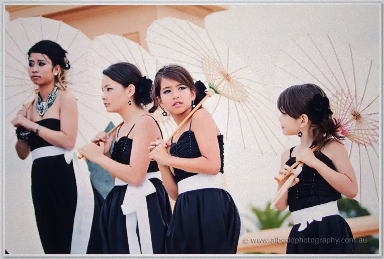 Fremantle Wedding | Charlene and Phil Bridesmaids and flower girls in black hold white paper parasols in a soft-lit ceremonial line