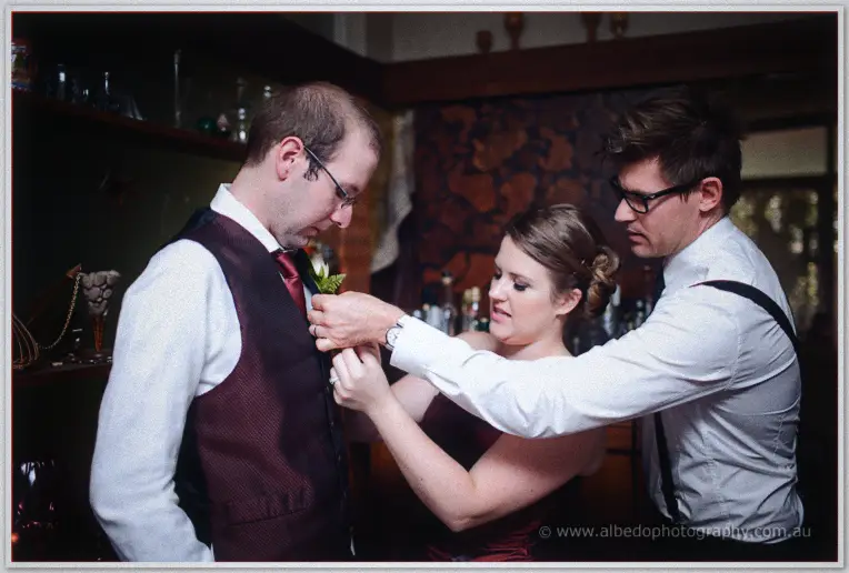 Attendants pin a green boutonniere to a man's burgundy vest in the timber bar before the ceremony
