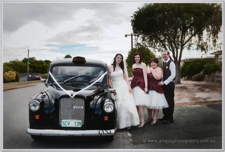 The bridal party poses beside a black vintage taxi with white ribbons across its grille on a suburban street