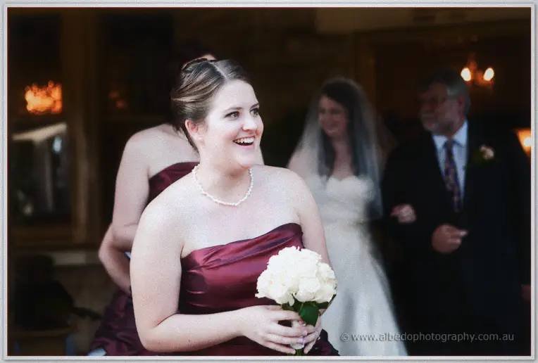 A beaming bridesmaid in burgundy satin carries white roses while Jenn follows behind in her veil