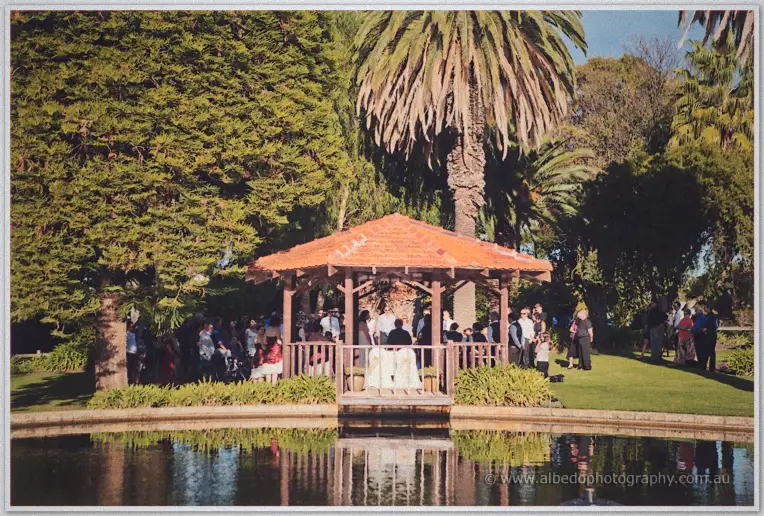 Guests gather under the terracotta-roofed gazebo on the lawn, its reflection stretched across the pond