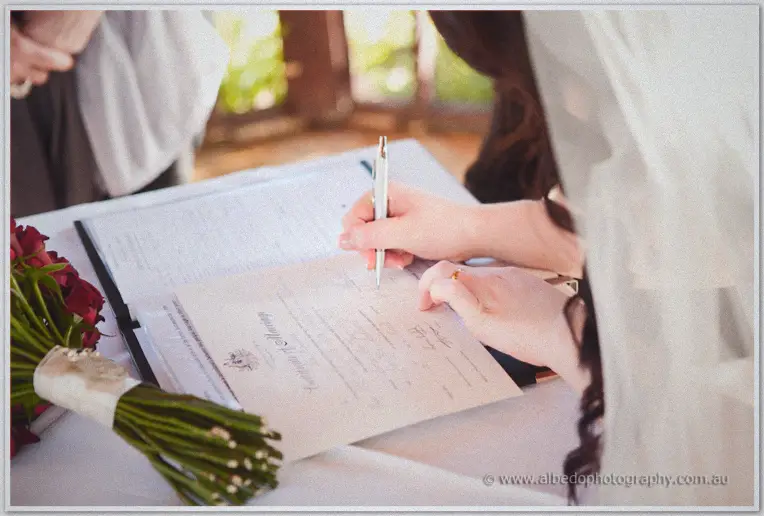 Jenn signs the marriage certificate with a white pen, red roses resting on the table beside her