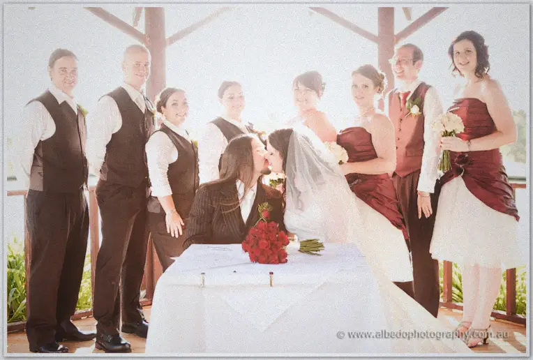 Jenn and Lance kiss over the signing table as their full bridal party gathers around in the gazebo light