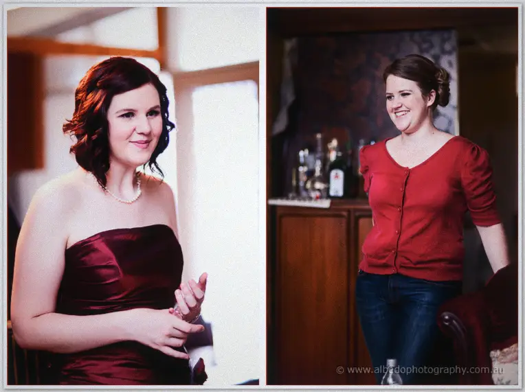 Two bridesmaids laugh while getting ready, one in a burgundy gown by the window and one in a red cardigan