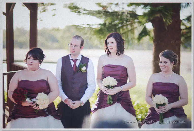 The bridal party waits under the gazebo, bridesmaids in burgundy holding white and red bouquets by the lake