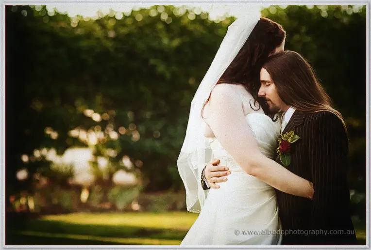 Jenn rests her head against Lance's long-haired shoulder in a sunlit garden, veil drifting in the breeze