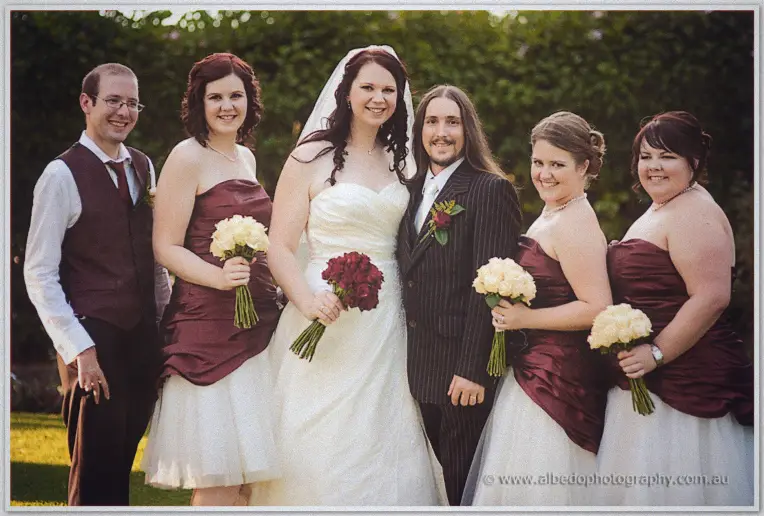 The full bridal party smiles on the lawn, bridesmaids in burgundy sashes holding cream and red bouquets