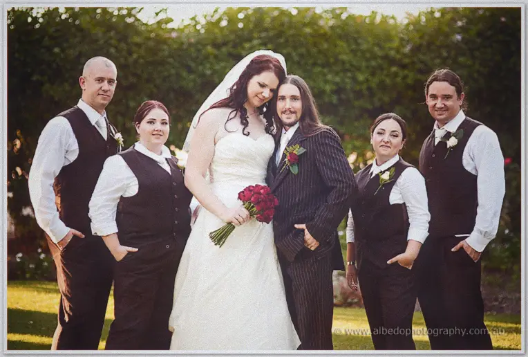 Jenn and Lance stand with four attendants in dark vests among the hedges in warm evening light