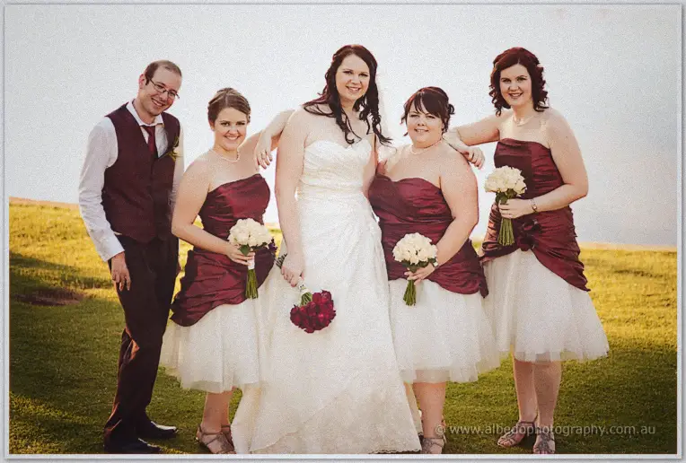 Jenn laughs with her bridesmaids on the sunlit hillside, arms linked, holding red and cream bouquets