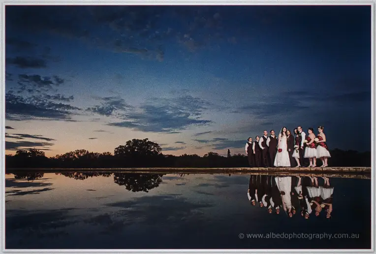 The whole wedding party lines the water's edge at twilight, doubled in the glassy pond below